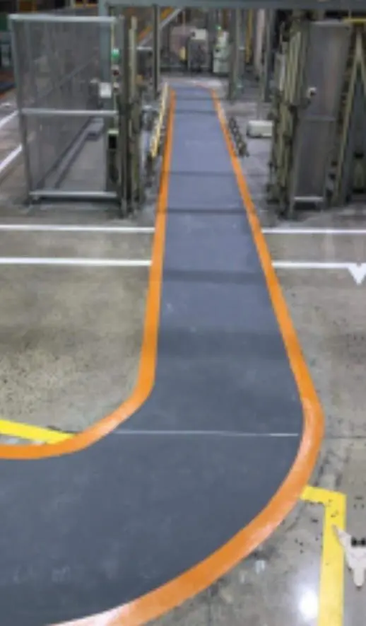 Pasillo de planta industrial con piso de poliuretano-cemento gris y demarcación de circulación en naranja siguiendo una curva